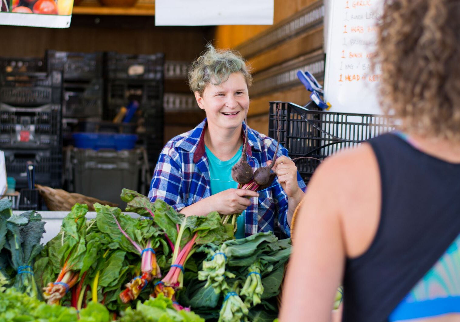 Smiling vendor at a market stand holding beets, talking to a customer in front of leafy greens.
