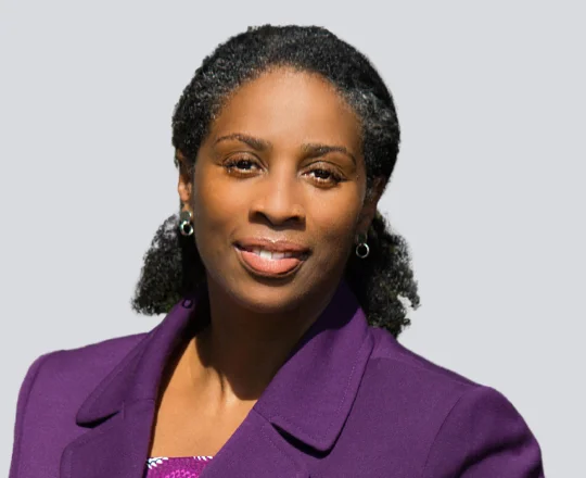 Woman in a purple blazer smiling, with short curly hair, against a plain light gray background.