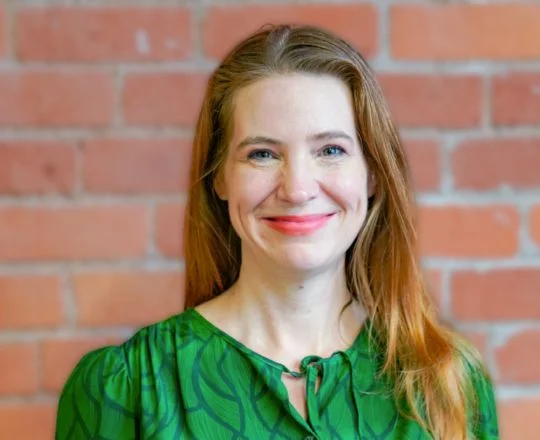 Woman with long red hair in a green blouse smiling, standing in front of a brick wall.