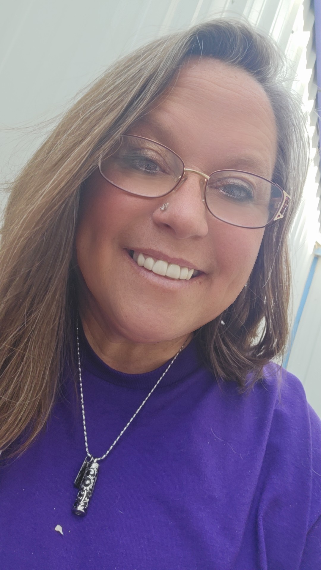 Smiling woman with glasses, long hair, and a purple shirt standing outside near a white building.