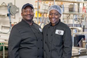 Two smiling chefs in black uniforms stand together in a commercial kitchen.