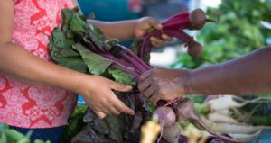 Two people exchanging fresh beets at an outdoor market, with green vegetables visible in the background.