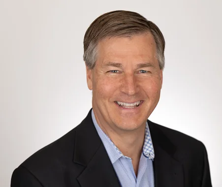 Smiling man in a suit jacket and blue shirt against a plain white background.