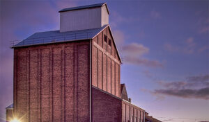A tall brick industrial building with a metal roof against a purple sky at sunset.