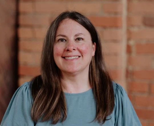 Woman with long brown hair, wearing a blue top, smiling in front of a brick wall background.