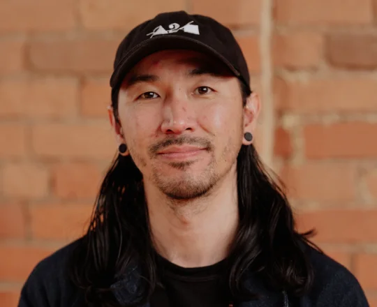 Person with long dark hair, a black cap, and earrings stands in front of a brick wall, smiling slightly.