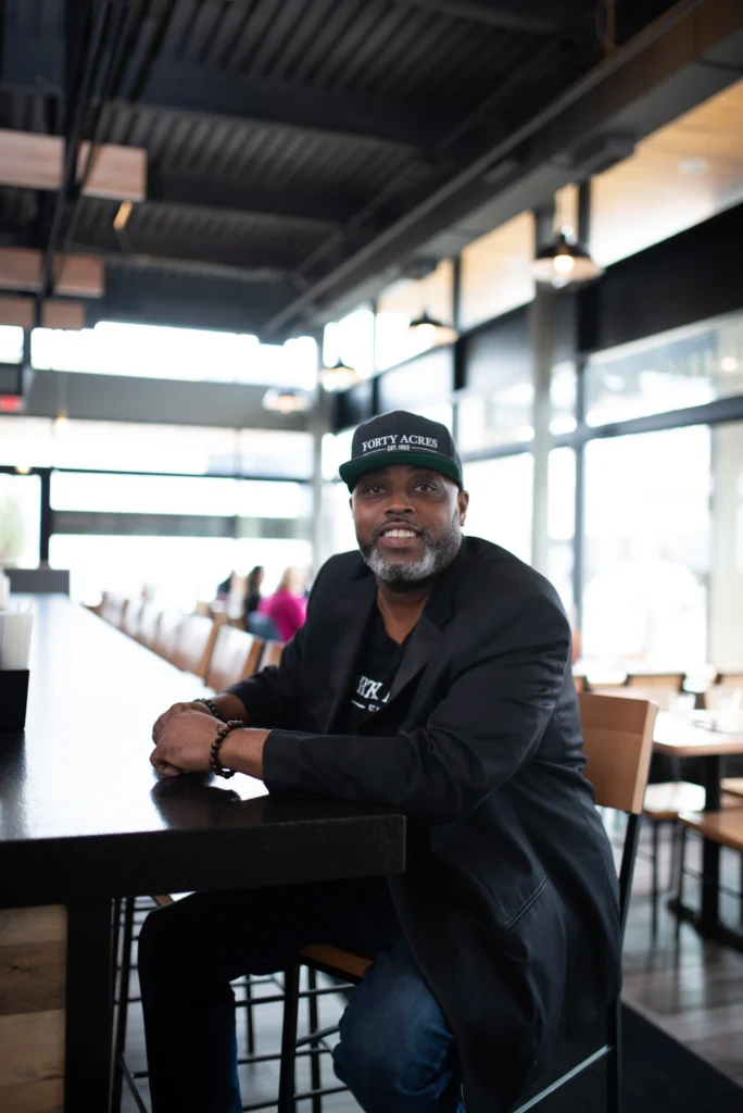 Man in a black jacket and cap sits at a bar in a bright, modern restaurant during the day.