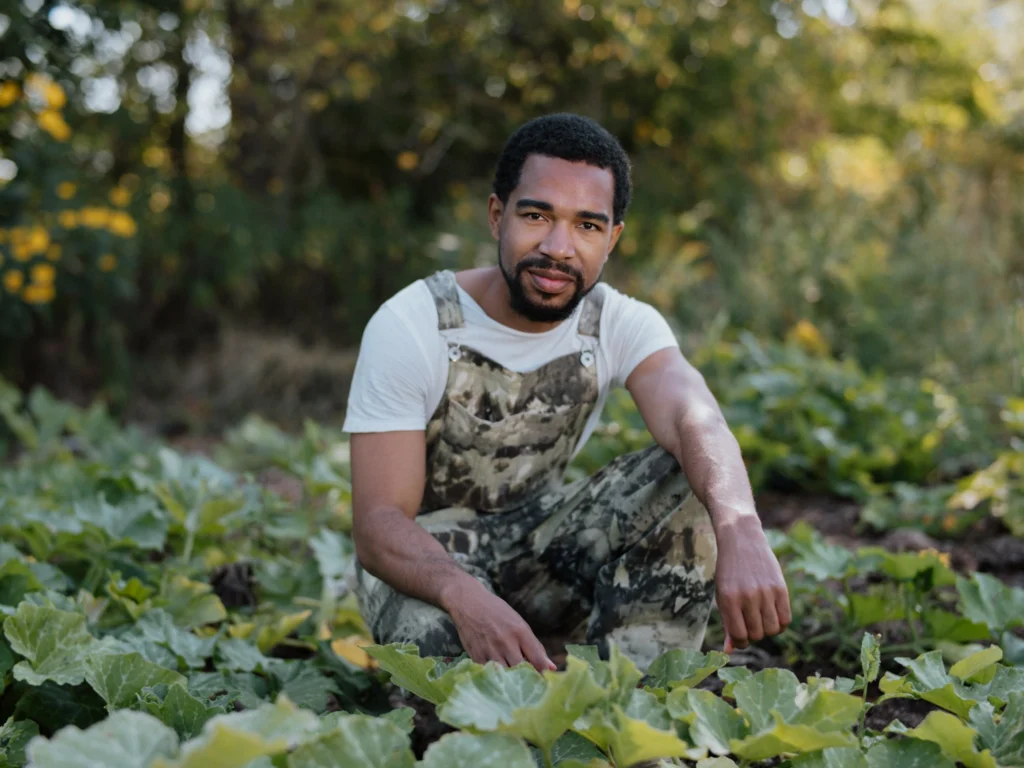 Man in camouflage overalls kneels in a leafy vegetable garden with trees in the background.