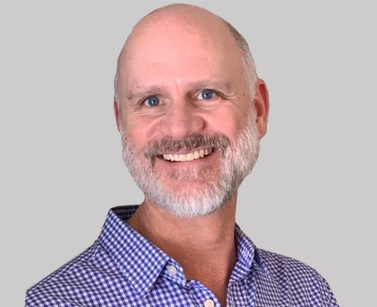 Smiling bearded man with a bald head in a blue checkered shirt, facing forward against a gray background.
