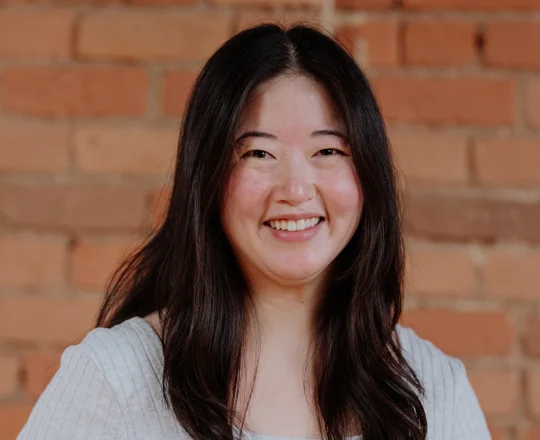 Woman with long dark hair smiling, wearing a light gray top, standing in front of a brick wall.