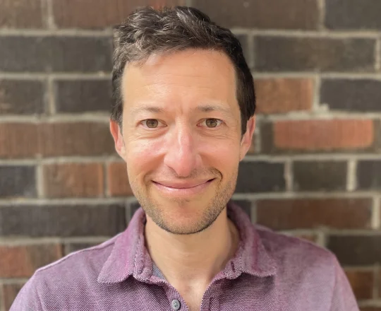 Smiling man with short brown hair and stubble, wearing a mauve polo shirt, standing in front of a brick wall.