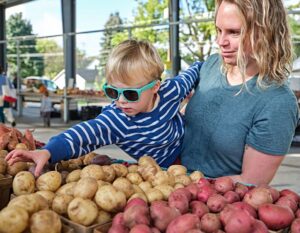Child in blue sunglasses reaches for potatoes at a market, held by an adult in a blue shirt.
