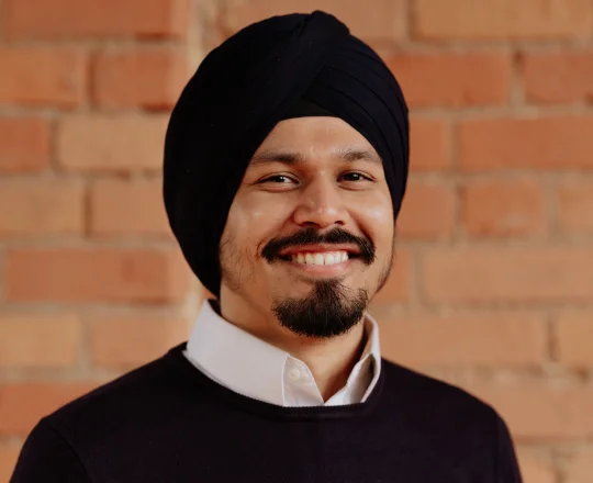 Man with a black turban and beard smiles at the camera, standing in front of a brick wall.