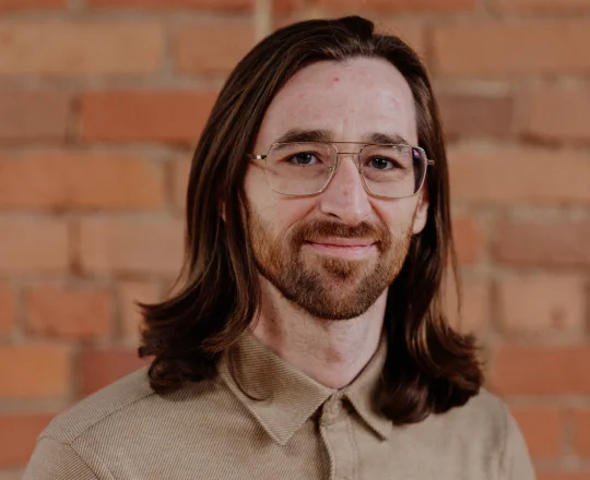 Man with long brown hair, glasses, and a beard smiling in front of a brick wall.