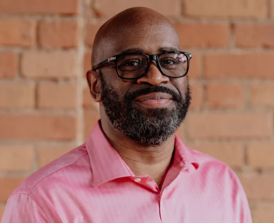 Man with a beard and glasses wearing a pink shirt stands in front of a brick wall, smiling slightly.