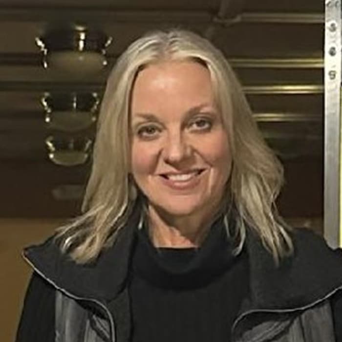 A woman with long blonde hair smiles while wearing a black jacket and standing indoors.