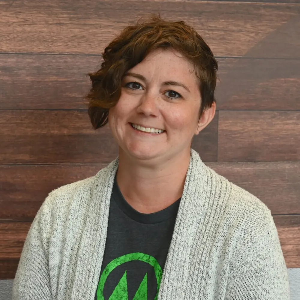 Smiling person with short brown hair, wearing a gray sweater and dark t-shirt, in front of wood wall.
