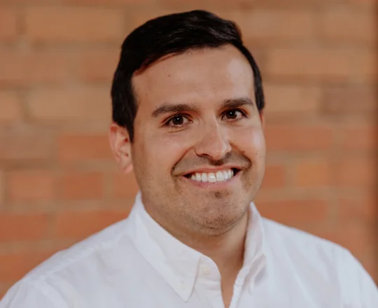 Smiling man with dark hair wearing a white shirt, standing in front of a brick wall background.