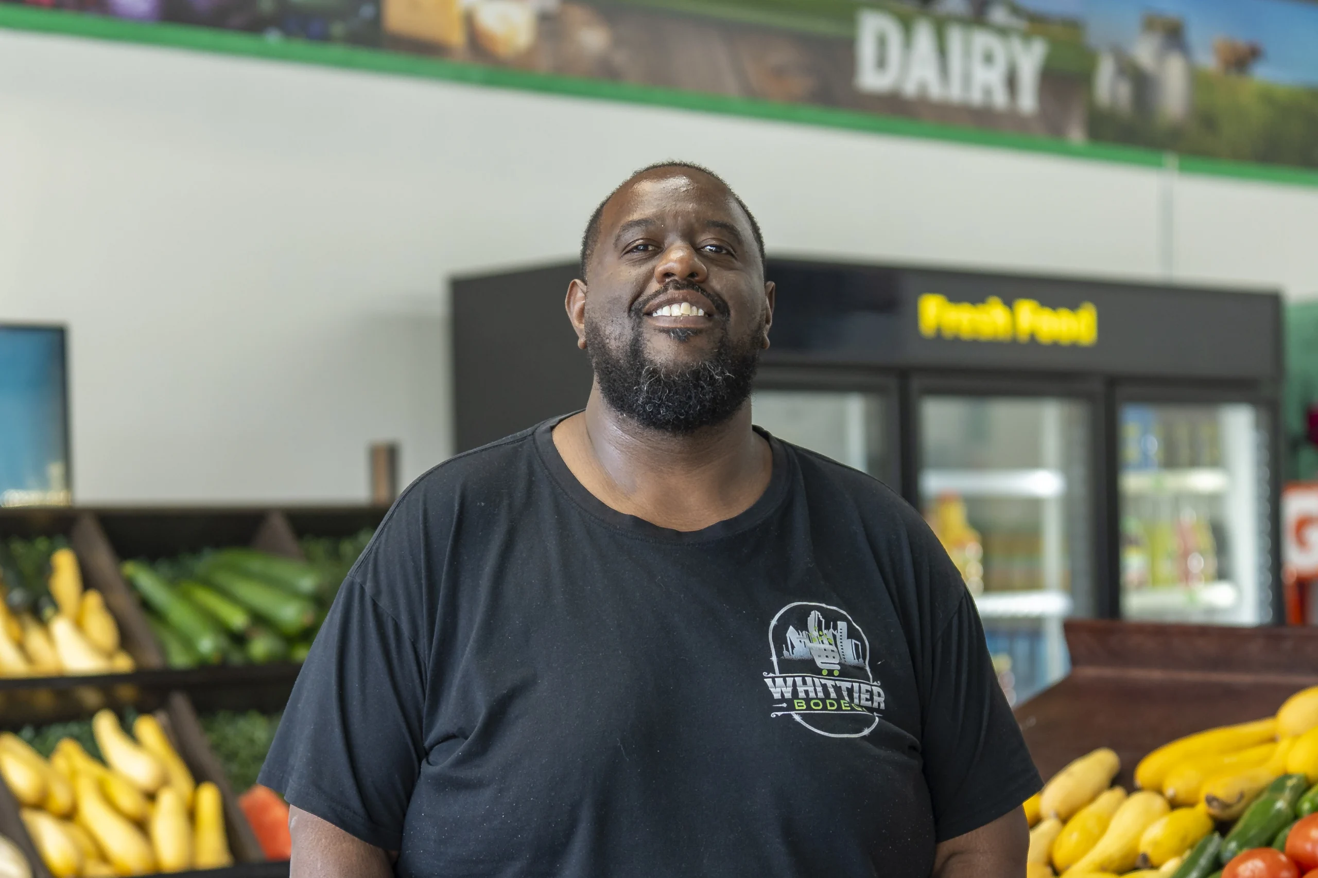 Man smiling in a grocery store produce section, wearing a black Whittier Boulder t-shirt.