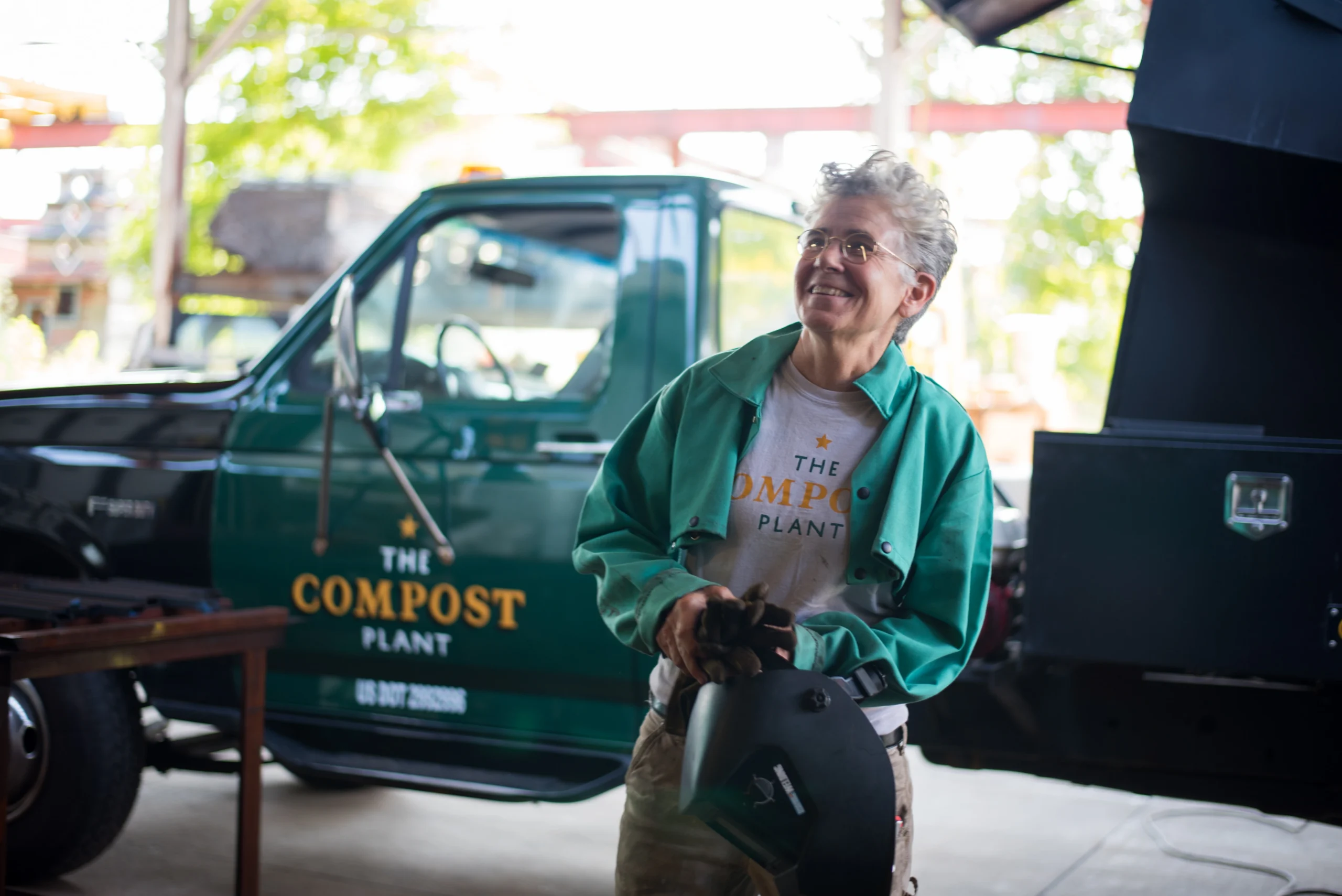 Smiling person in a green jacket holds a welding mask near a truck labeled The Compost Plant..