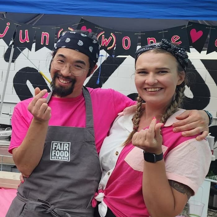 Two people smiling in pink outfits, making finger hearts, standing under a banner at an outdoor event.