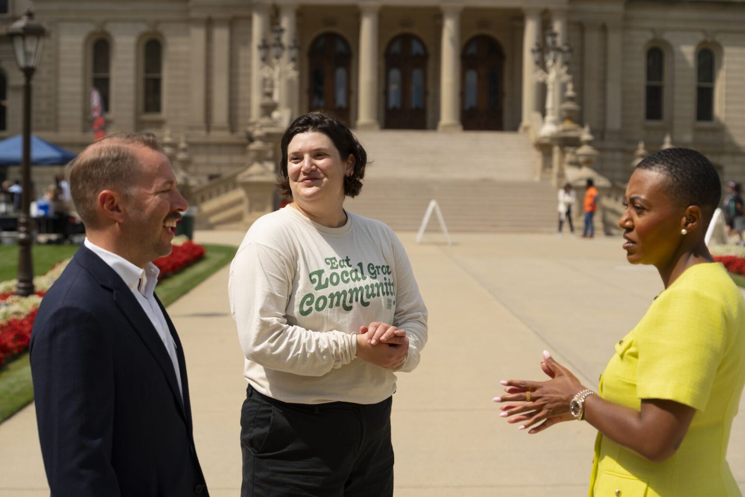 Three people talk outside a large, ornate building on a sunny day. One wears a Local Green Community shirt.