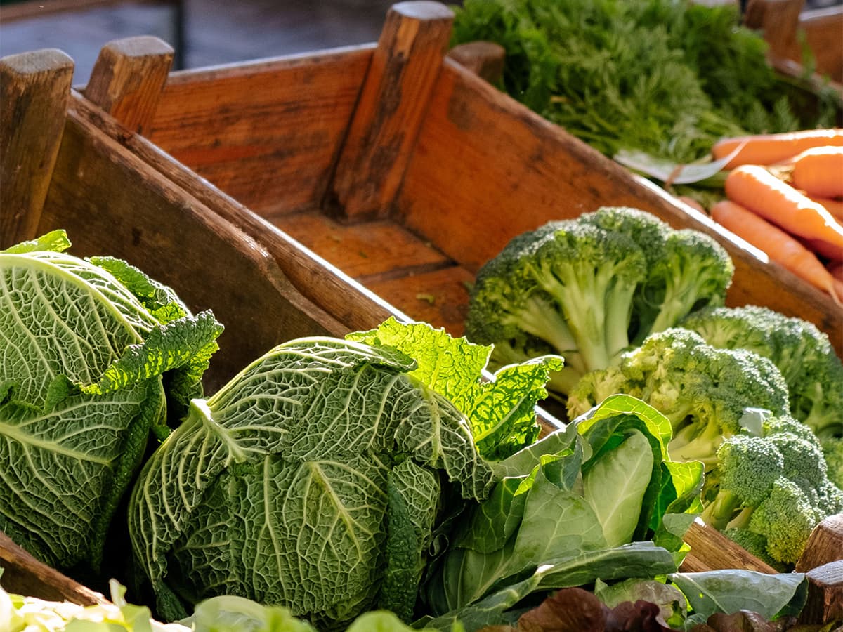 Green cabbage, broccoli, and carrots displayed in wooden crates at a fresh produce market.