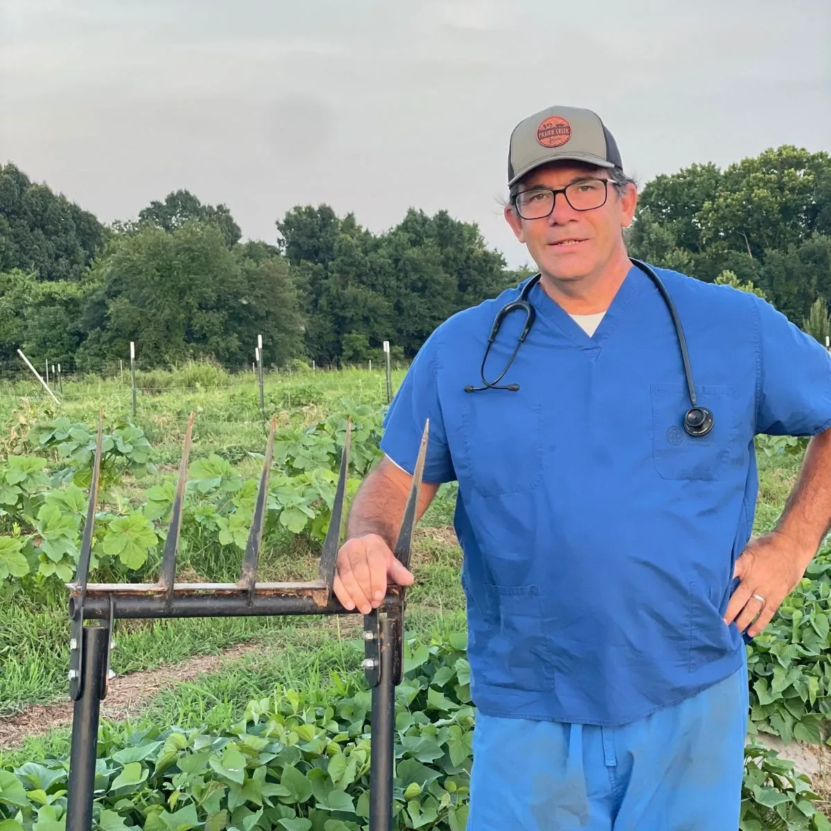 A man in blue scrubs with a stethoscope stands in a garden, leaning on a pitchfork, with green plants behind him.