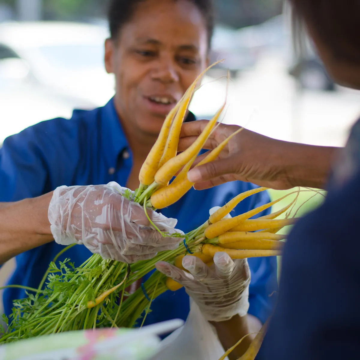 A person wearing gloves hands a bunch of yellow carrots to another person at an outdoor market.