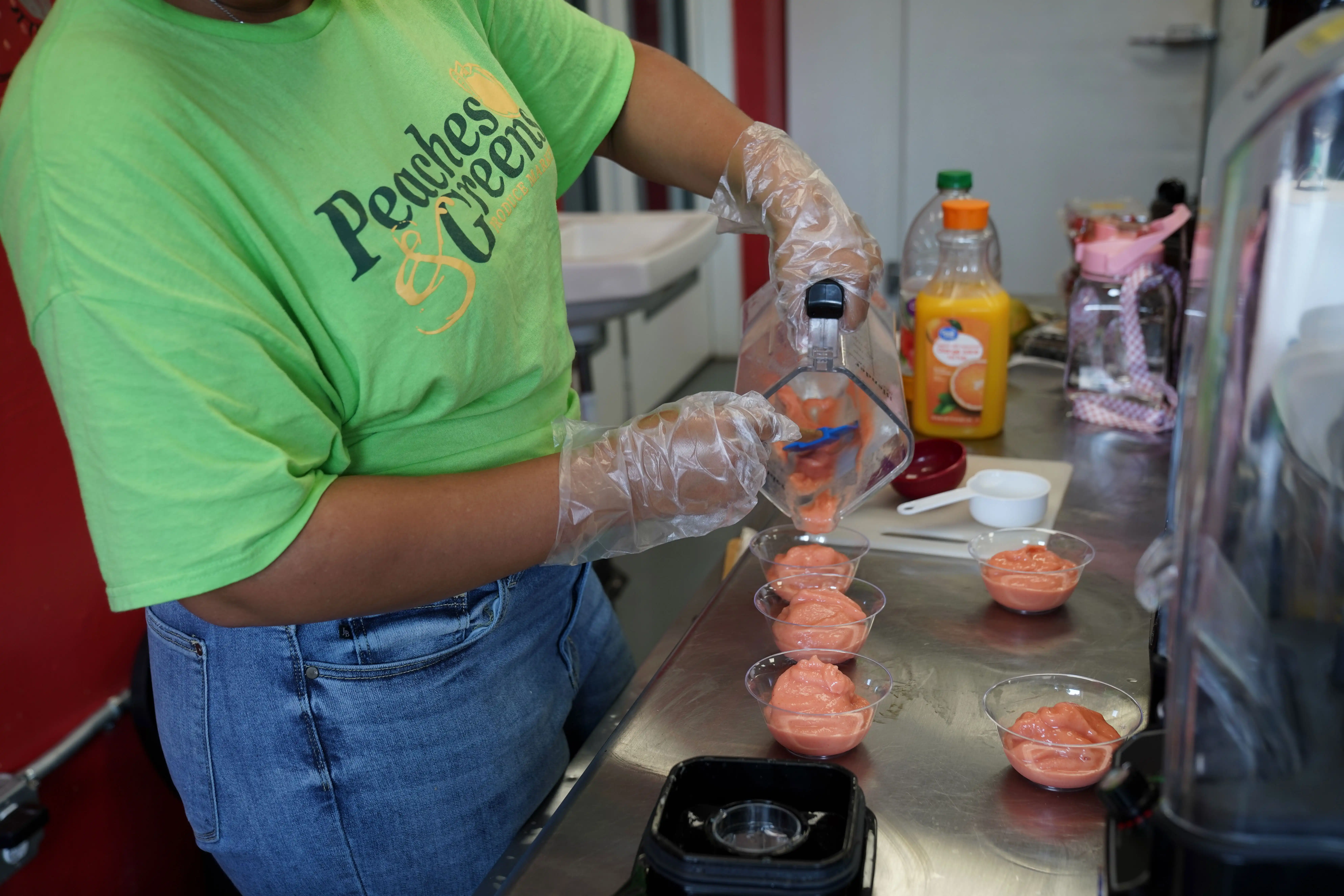 Person in gloves pours pink smoothie into cups, wearing a green Peaches & Greens shirt near juice bottles.