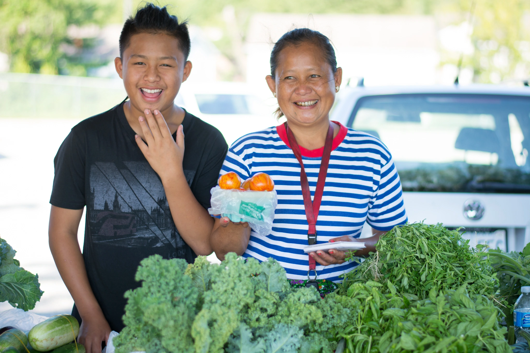 Smiling woman holding tomatoes and boy standing behind a table of leafy greens at an outdoor market.