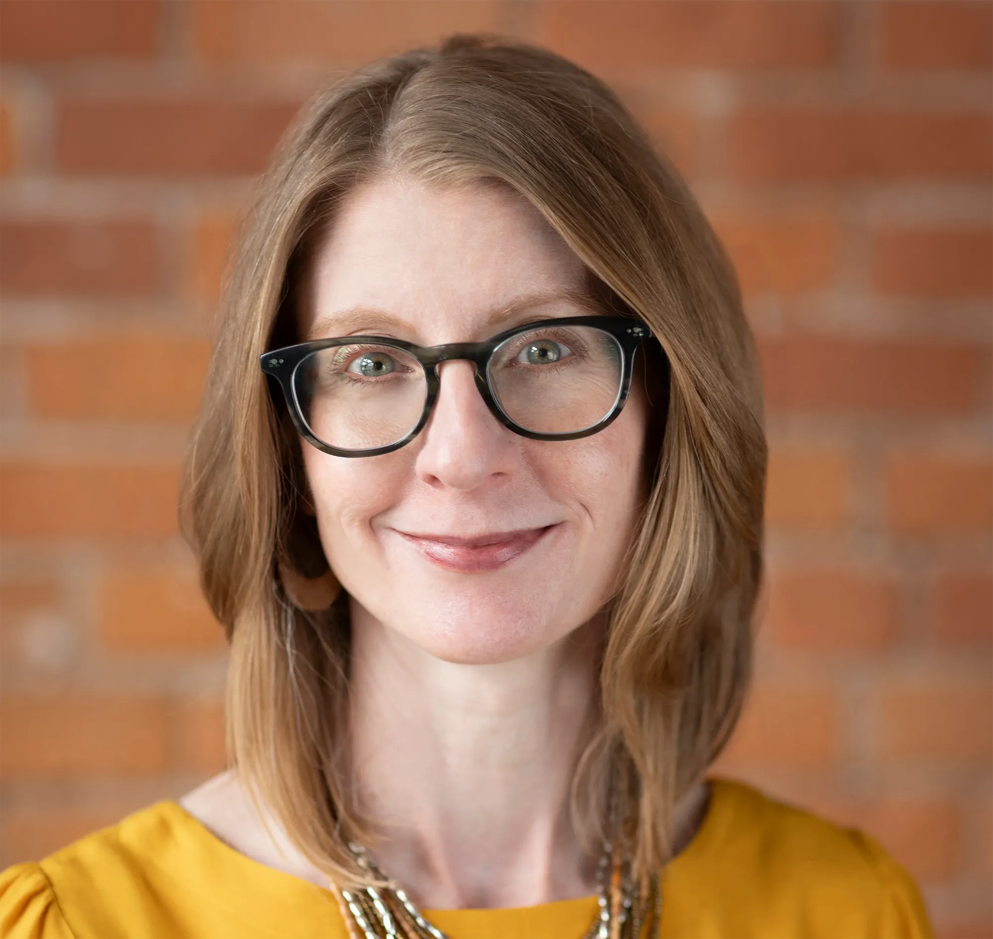 Woman with glasses and shoulder-length hair, smiling, wearing a yellow top, with a brick wall background.