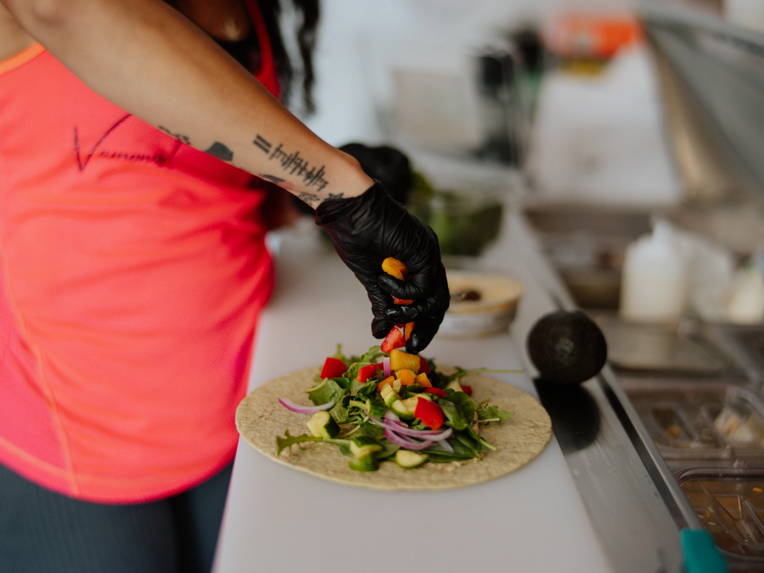Person in a pink shirt adds veggies to a tortilla wrap while wearing black gloves at a prep counter.