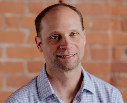Smiling man with short hair in a plaid shirt stands in front of a brick wall.