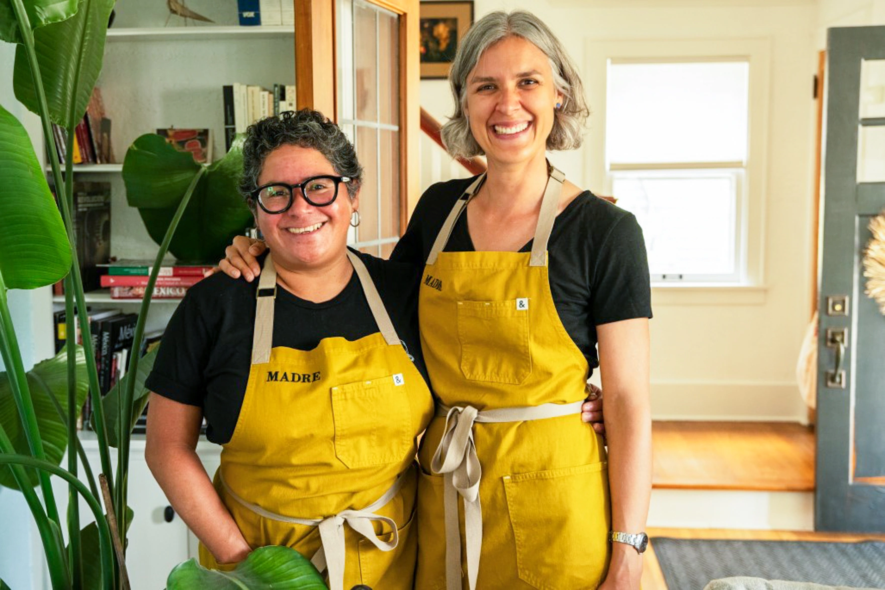 Two women in yellow aprons smile and pose together in a cozy, sunlit kitchen.