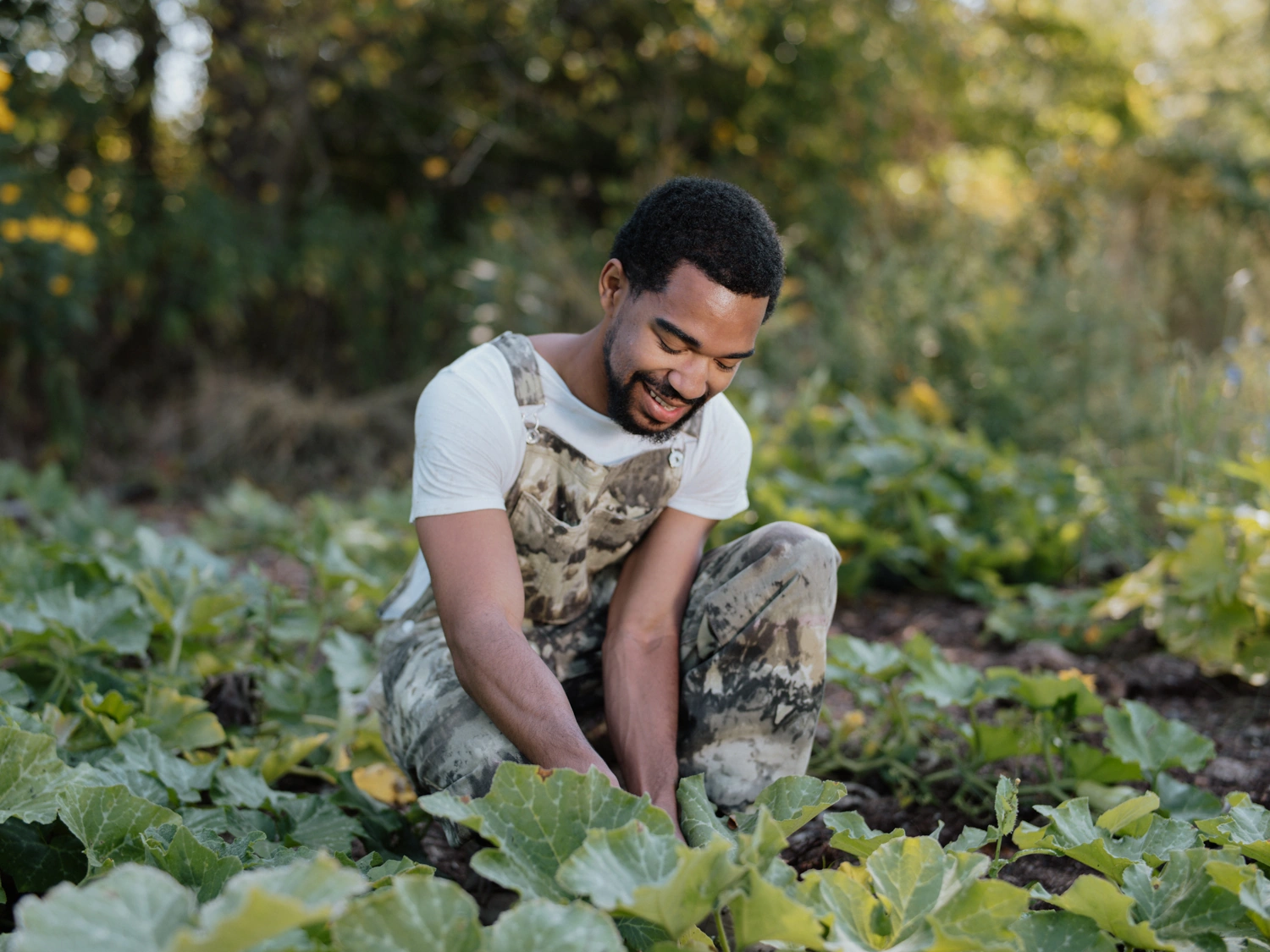 Man in camo overalls smiles while gardening outdoors, surrounded by green leafy plants.