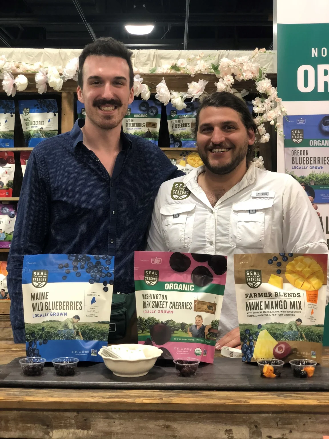 Two smiling men stand behind a table displaying bags of dried fruit and small samples at a booth.