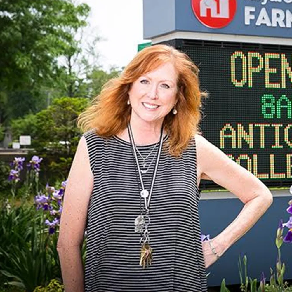Woman with red hair in striped dress stands outside near flowers and a digital sign reading OPEN.