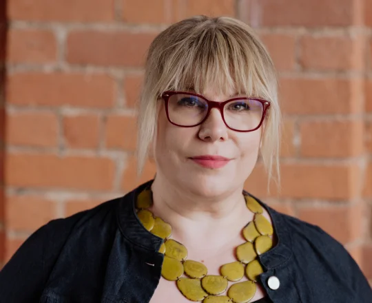 Woman with blond hair, red glasses, and yellow necklace standing in front of a brick wall.