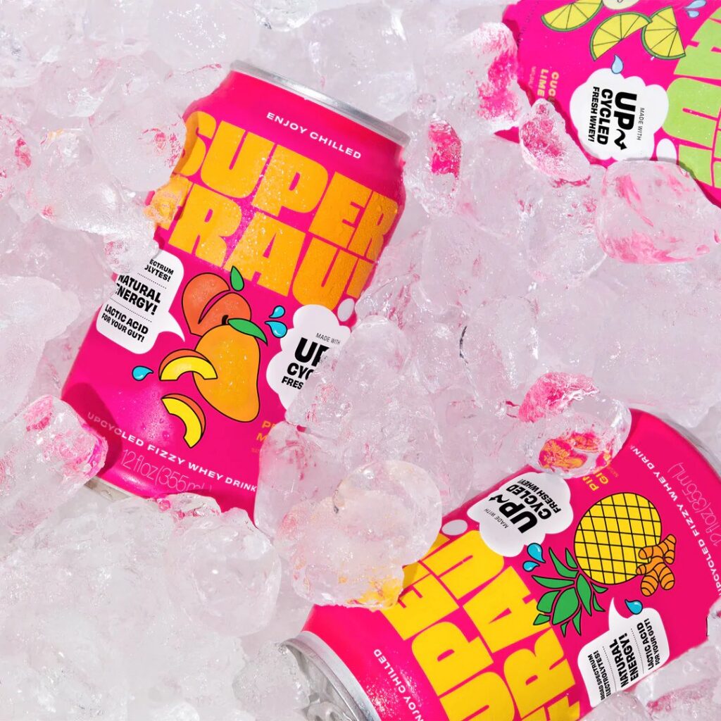 Pink Superfrau cans with fruit illustrations are chilled on ice, partially covered by ice cubes.