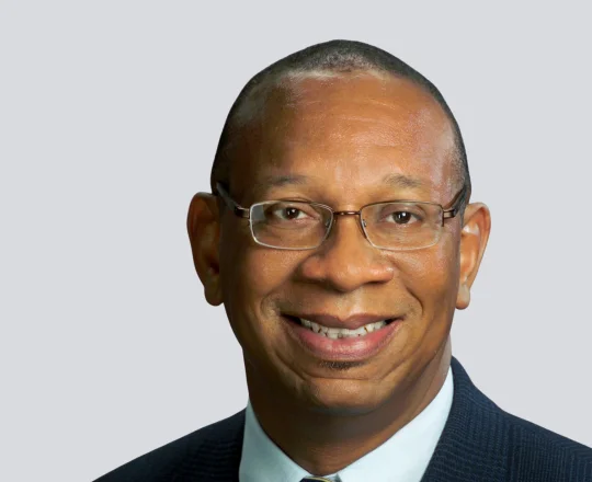 Smiling man wearing glasses, a suit, and tie, pictured against a plain light gray background.